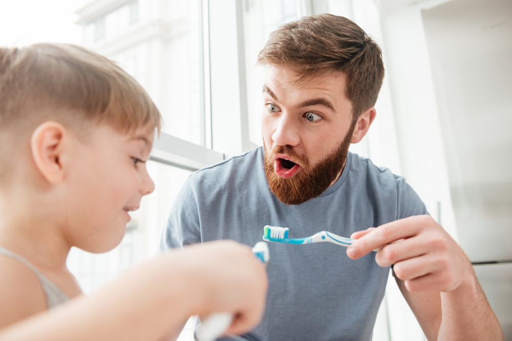 Father and son smiling while brushing teeth in bathroom. - Best Dental Care: 6 Tips for Having a Healthy Mouth